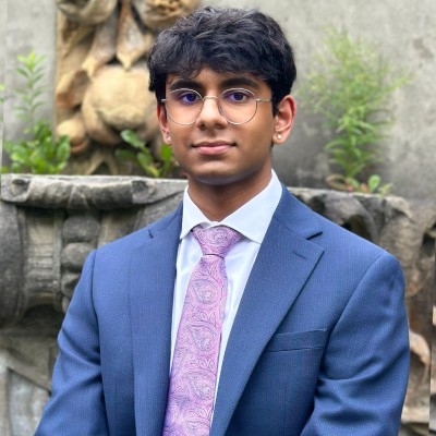 A square photo of Anant Dhokia wearing round silver glasses and a blue suit jacket with a pink tie and white button up shirt.