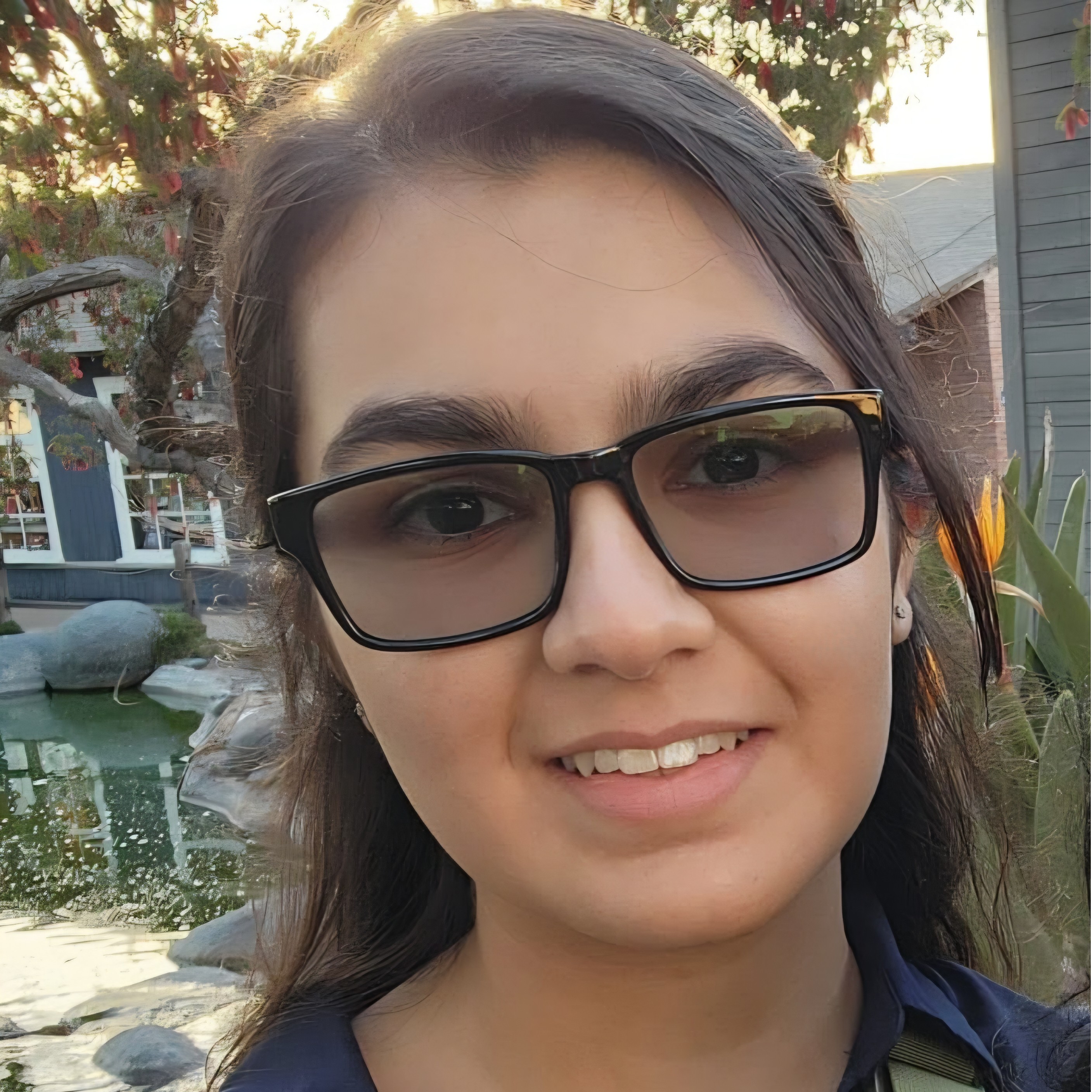Smiling person with long hair and glasses, wearing a collared shirt. Trees and a pond are visible in the background.