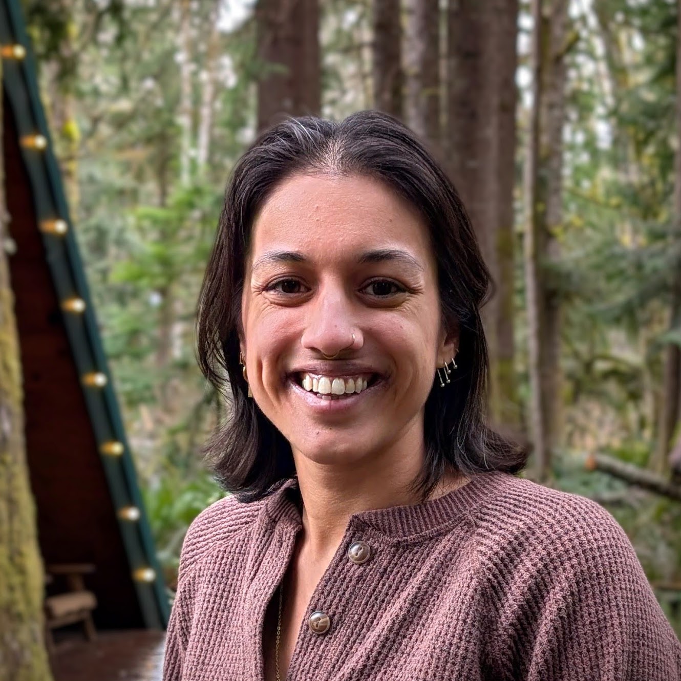 Anjali Pal, a half-Asian woman, smiles directly at us, with shoulder length dark brown hair, standing outside in front of some trees, wearing a brown sweater.