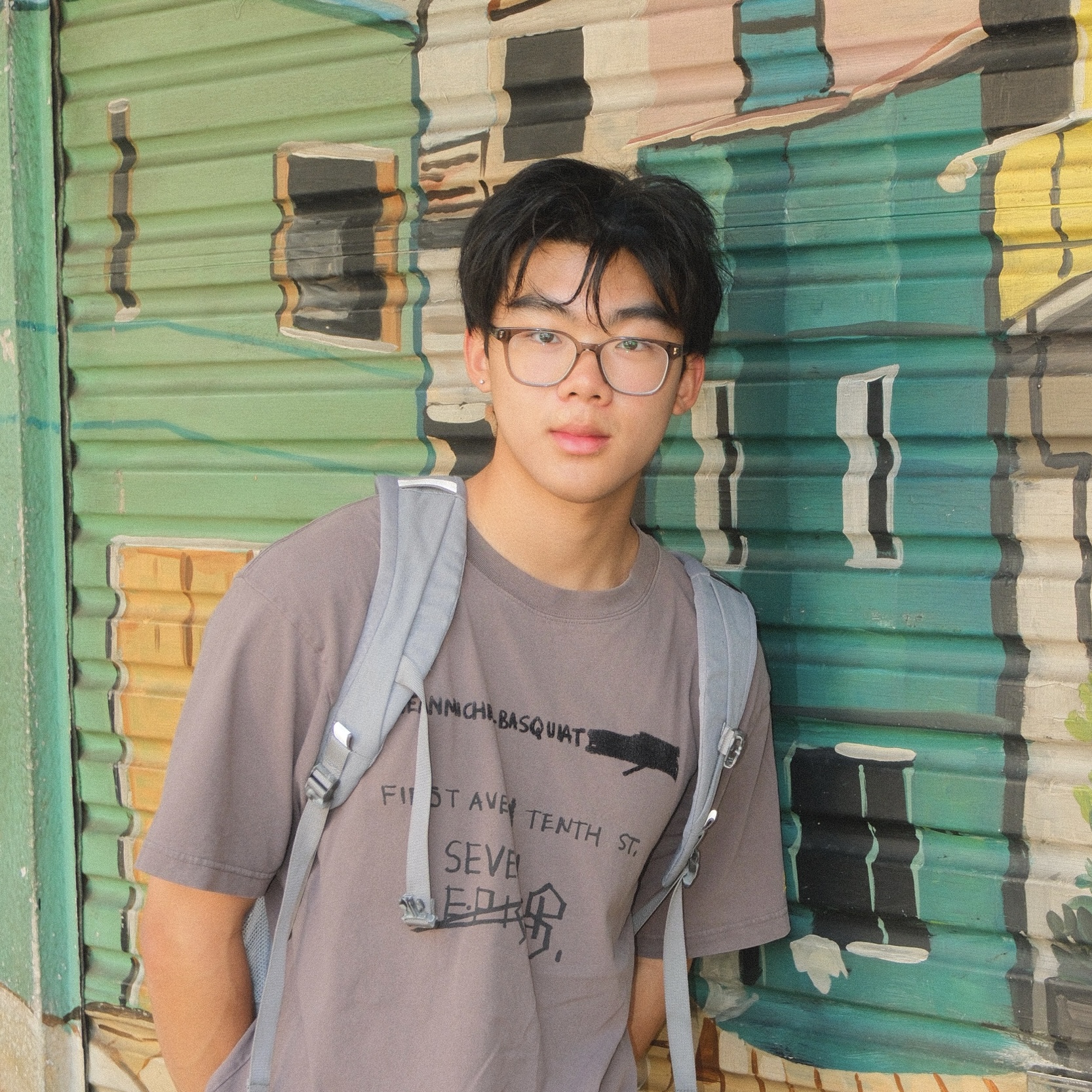 Avery wearing glasses and a backpack in front of a colorful mural, smiling at the camera.