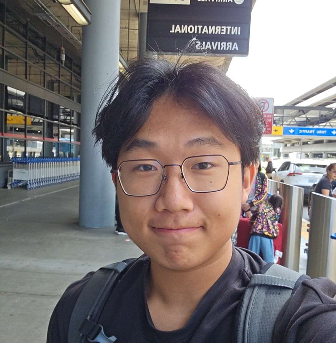 A selfie of a man with middle part black hair, glasses, and standing in front of an airport's arrivals area.