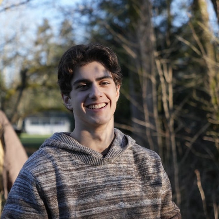 Headshot of Jamie, a white man with brown hair,  smiling at the camera in front of trees, wearing a brown sweater.