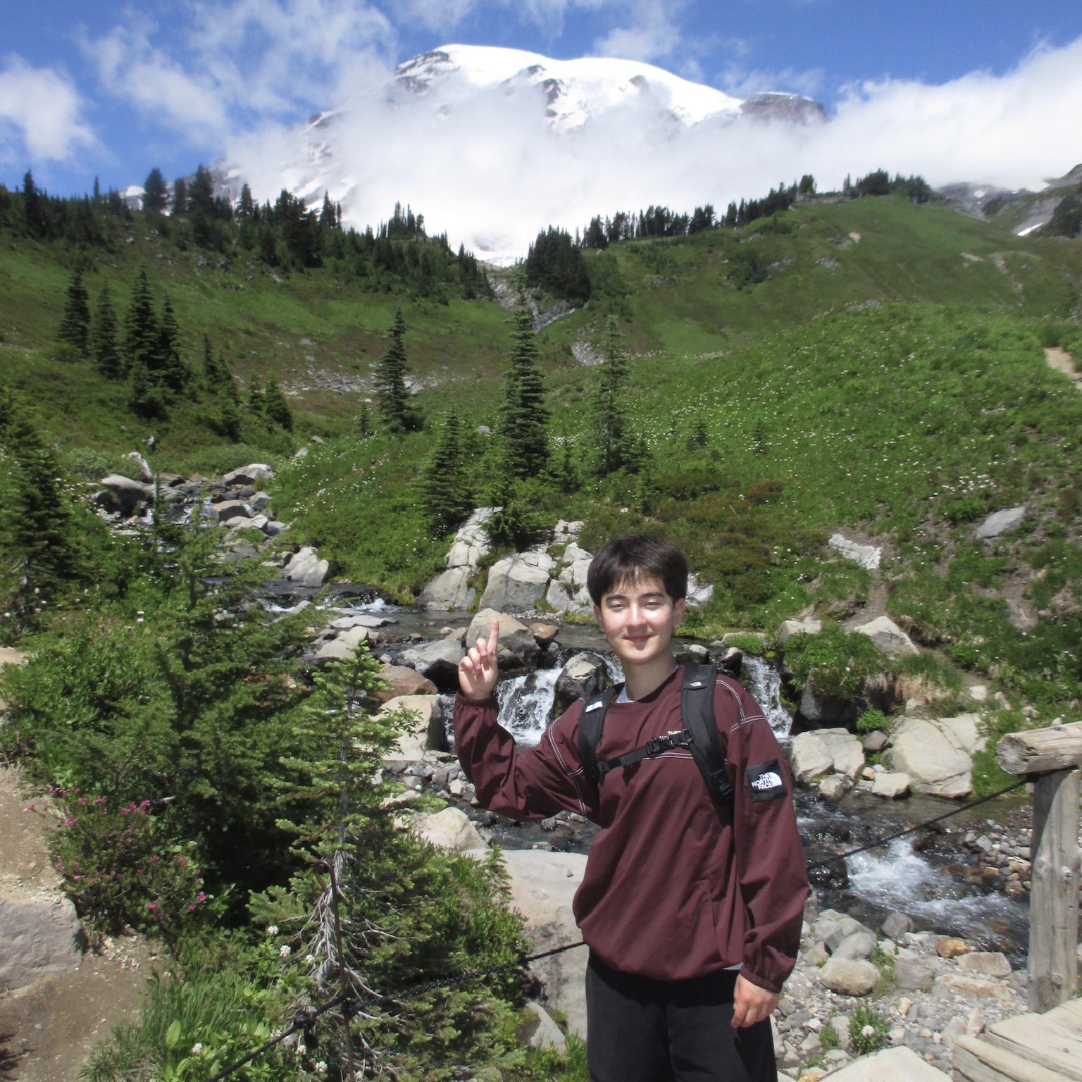 Lucas on a hiking trail, pointing at Mount Rainier in the background while smiling at the camera.