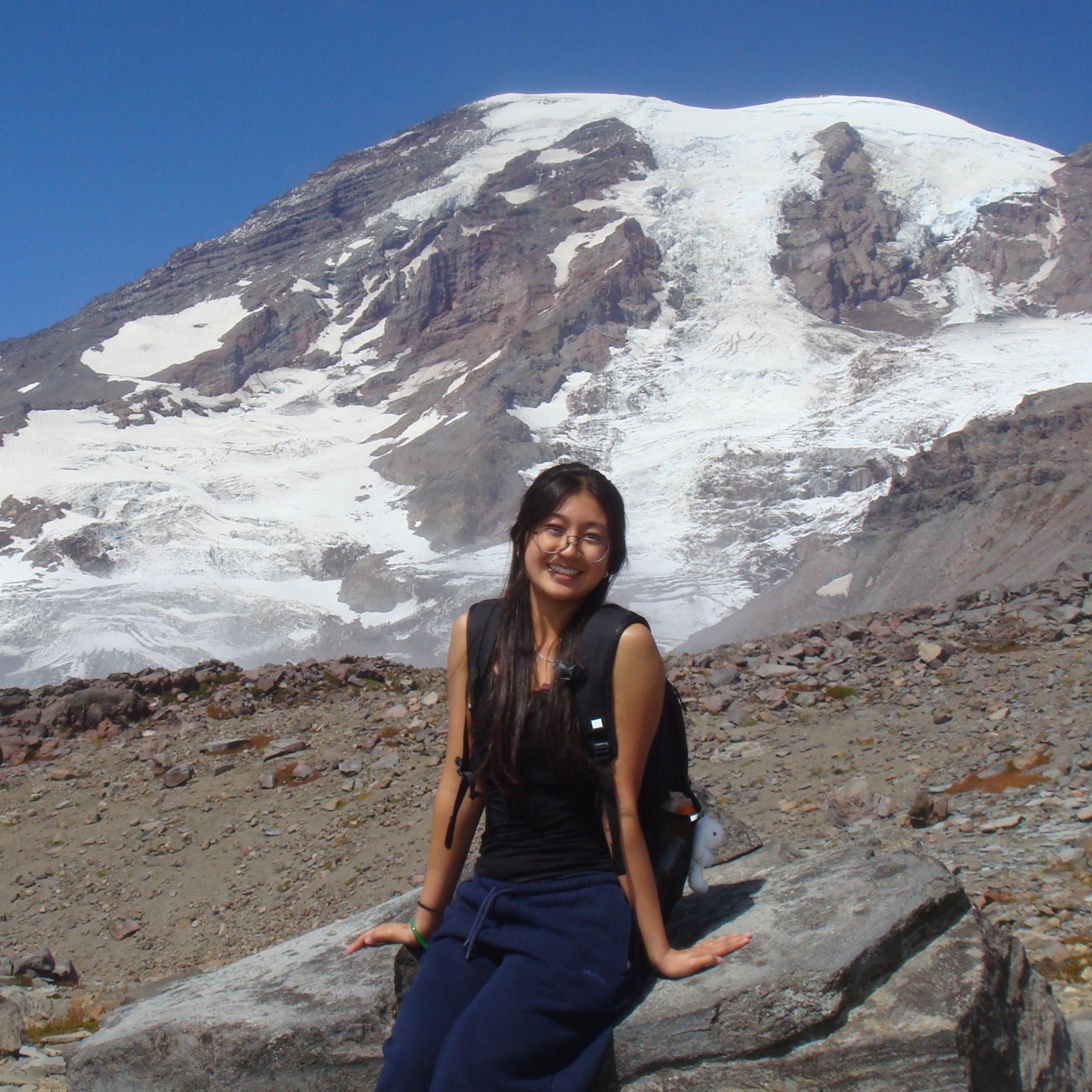 A woman sitting on a large rock with a backpack, smiling in front of snow-covered Mount Rainier under a clear blue sky