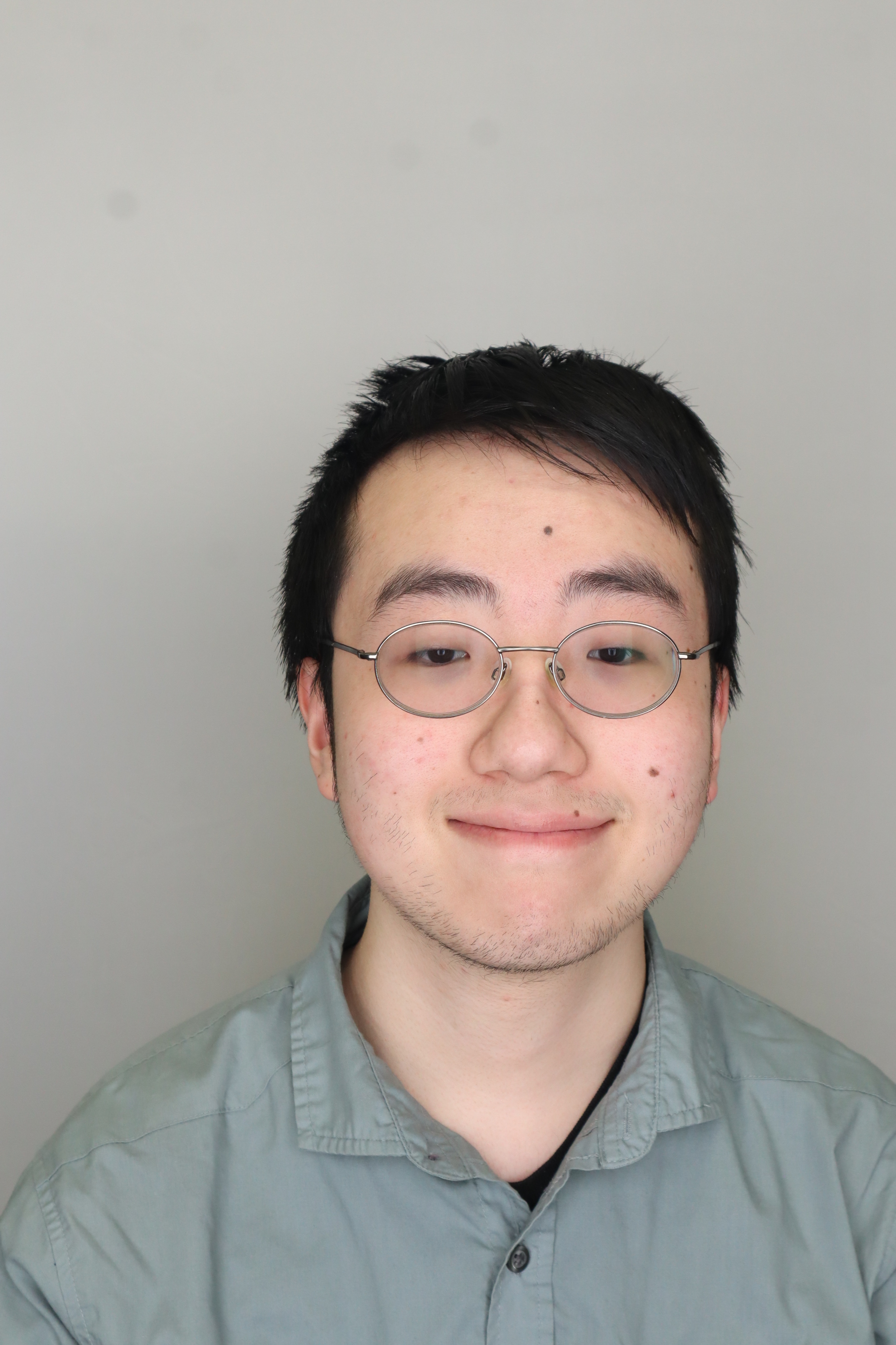 A headshot of Ethan Li standing in front of a grey background and smiling, he is an Asian man with black hair and glasses.