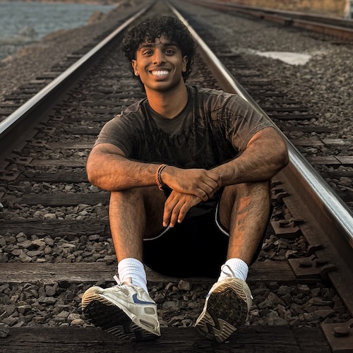 South Asian man with dark curly hair and a wide smile sits cross-legged on railroad tracks at sunset. He wears a dark graphic tee and white sneakers behind a background of water and light from sunset.