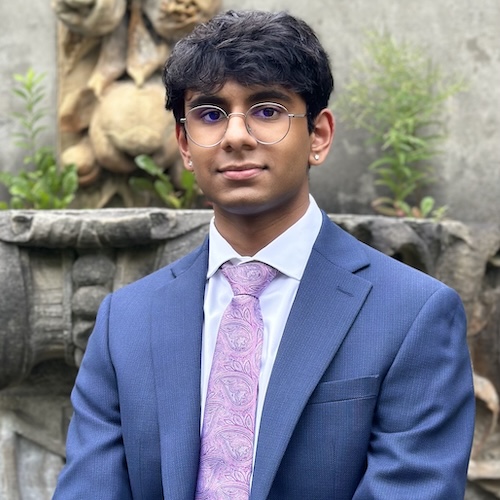 Anant is a south asian male with black hair and round, silver glasses. He's wearing a dark blue suit with a white button up shirt and a purple tie with swirling leaf-like designs on it. The blurred background shows a stone wall with some green vegetation.
