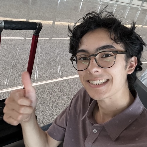 Hayden smiling and giving a thumbs up! He's sitting next to his suitcase in the airport.