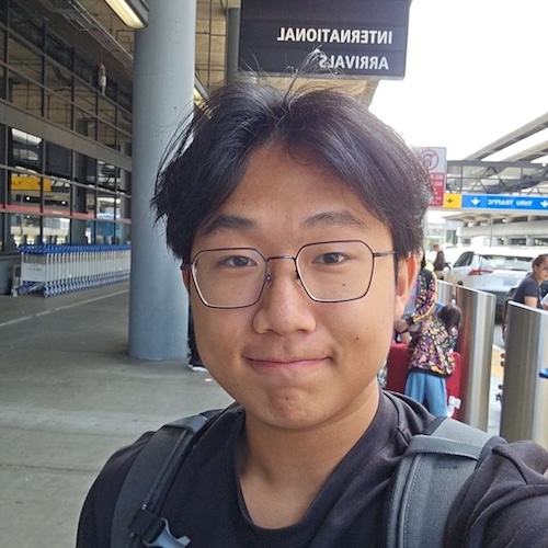 A selfie of an Asian man with glasses and a middle-part hairstyle in a black T-shirt in front of an airport terminal.