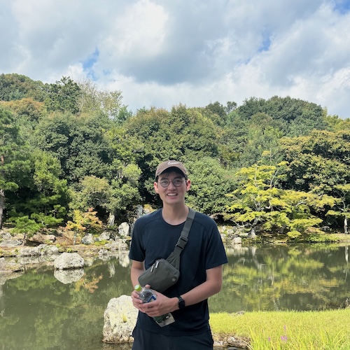 Spencer is standing in front of a garden. He wears glasses and is wearing a blue shirt and is smiling.