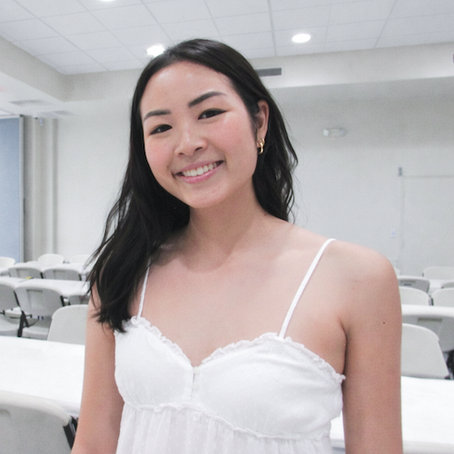 Smiling woman with shoulder-length dark hair wearing a white top, standing in a bright banquet room with rows of empty chairs in the background.