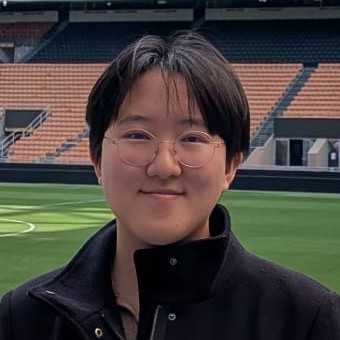 Cayden facing the camera, smiling slightly against a background of green grass and orange chairs. Cayden is East Asian with short black hair and is wearing a black coat and tan sweater.