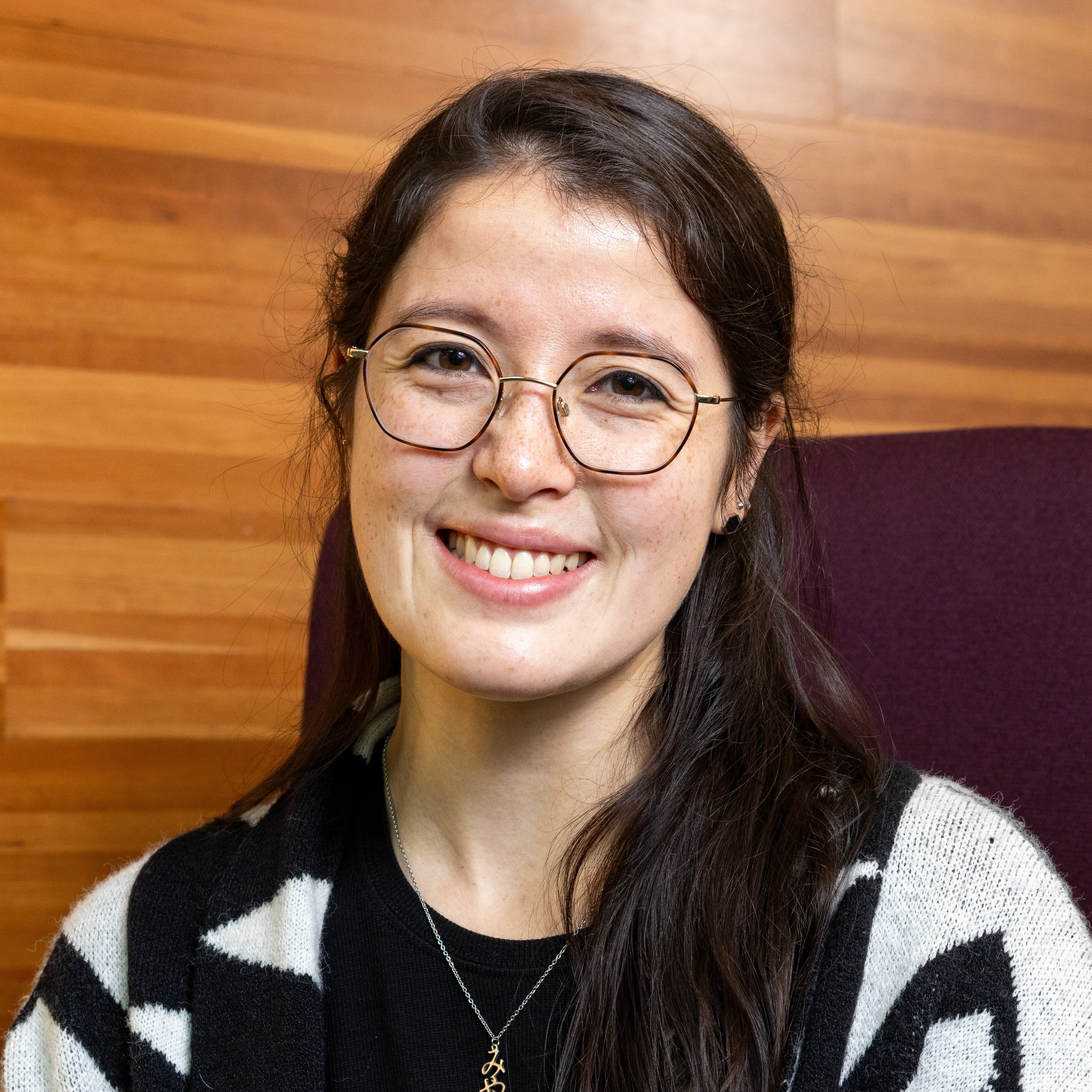 Miya Natsuhara, a half-Asian woman, smiles directly at us, with long dark brown hair, leaning against a wood-paneled wall, wearing a black and white sweater with geometric pattern.