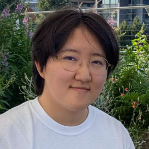 A headshot of Cayden facing the camera, smiling slightly against a background of greenery. He is East Asian with short black hair and is wearing a white T-shirt.