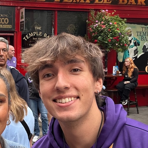 Dalton is a lighter skinned man with brown hair coming down to his eyebrows. He is wearing a purple sweatshirt and is smiling.