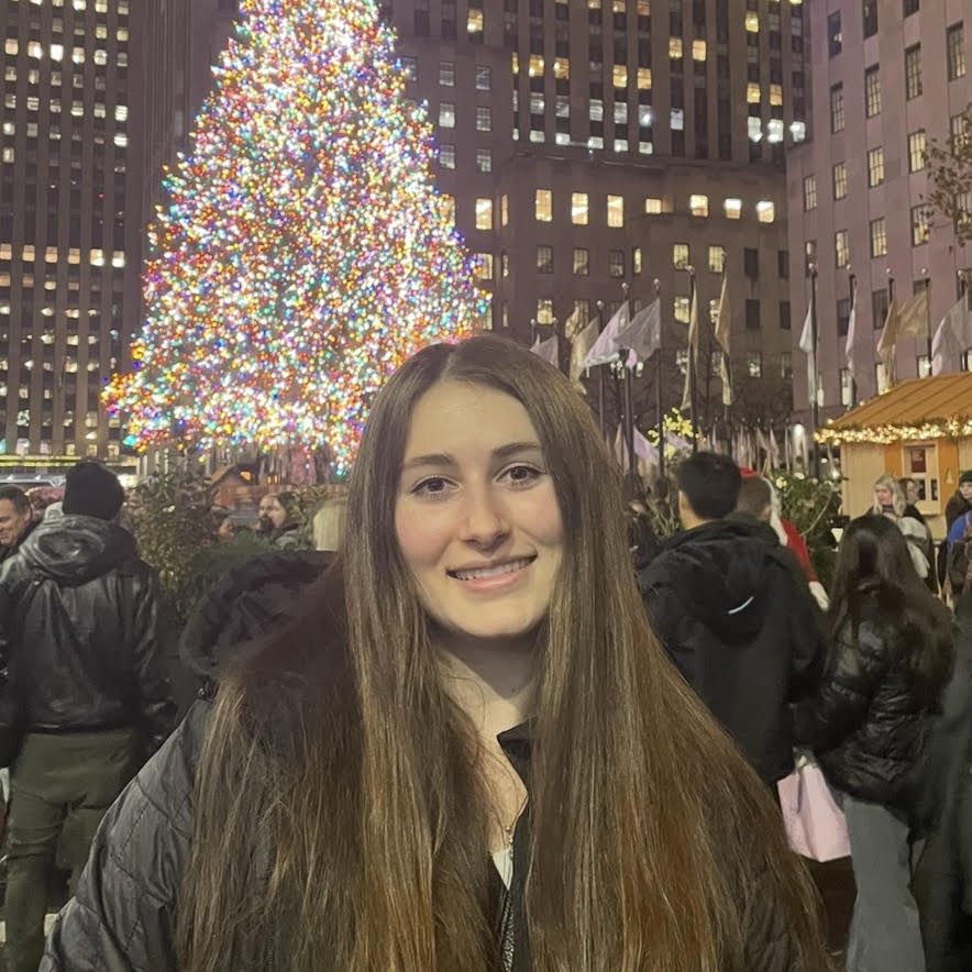 Hannah Swoffer smiles directly at us, with long dark brown hair, posing in front of the Christmas tree at Rockefeller Center