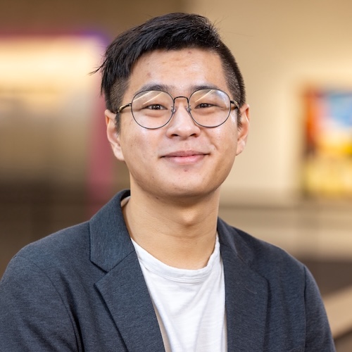 Matt Wang, an Asian man, smiles directly at us, with short black hair. He is against a gray background with white spots, wearing a white t-shirt with a blue jacket open.