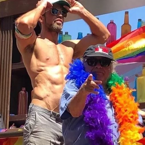 Danny Devito at a Gay Pride Parade, wearing a rainbow feather boa and looking happy.