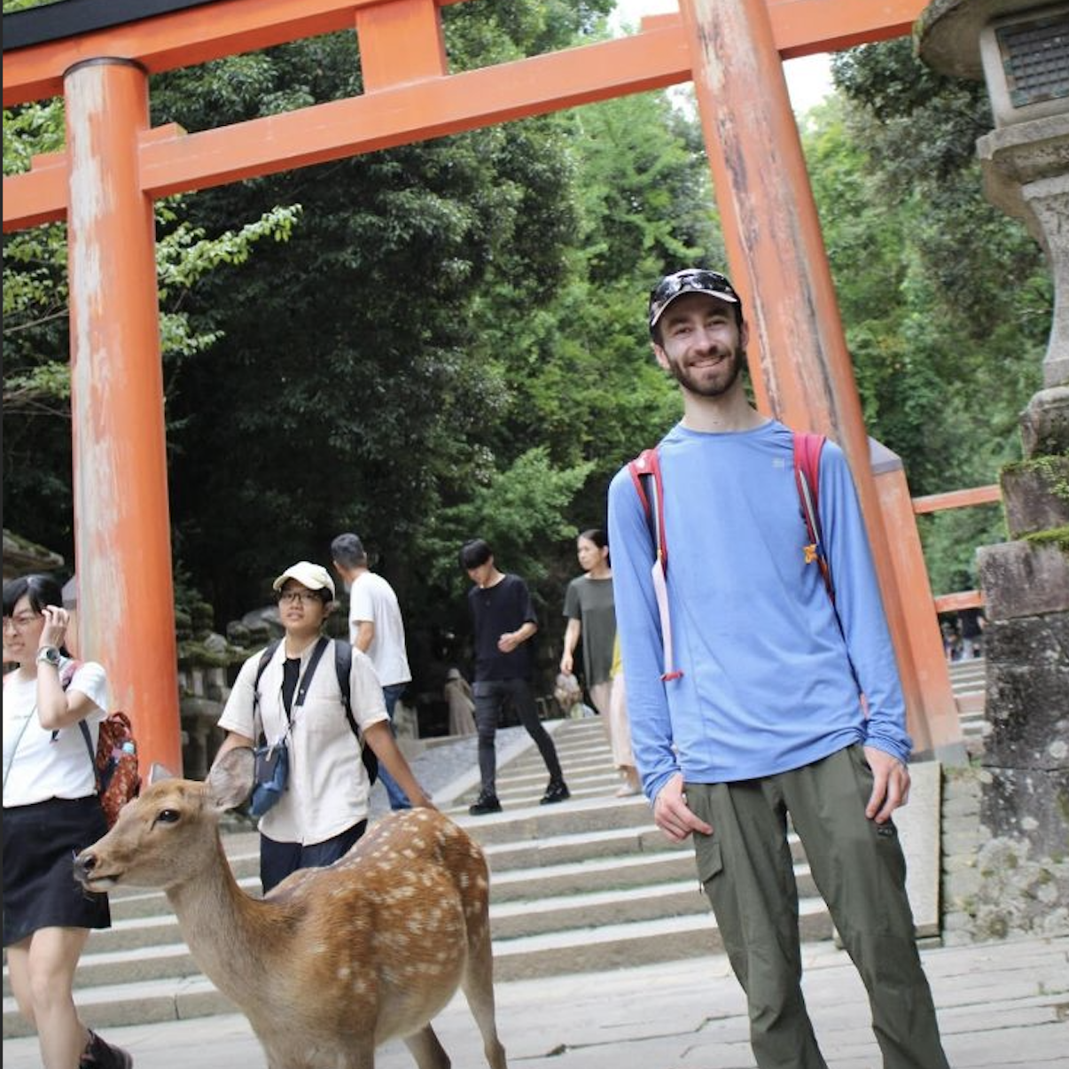 A profile picture of Adam Fuegmann smiling at the camera while
            standing beside a deer in Nara, Japan. He is a white man wearing a baseball cap, a blue
            sun shirt, and green khaki pants.