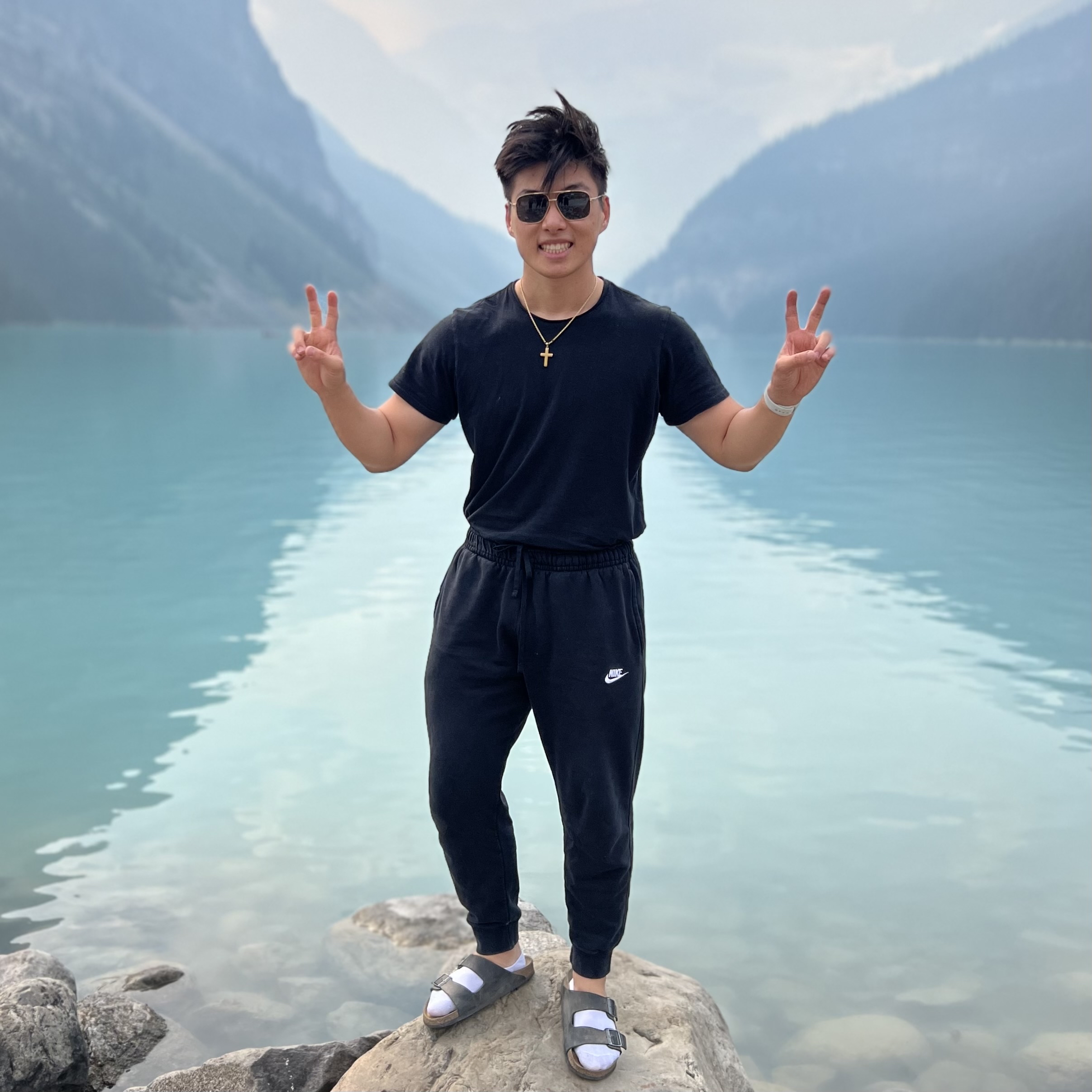 A profile picture of Alex Zhang, a young man standing on a rock at
            the edge of a turquoise lake with mountains in the background. He has short black hair
            and is smiling while wearing sunglasses. He is dressed in a black t-shirt, black Nike
            sweatpants, and gray Birkenstock sandals with white socks.