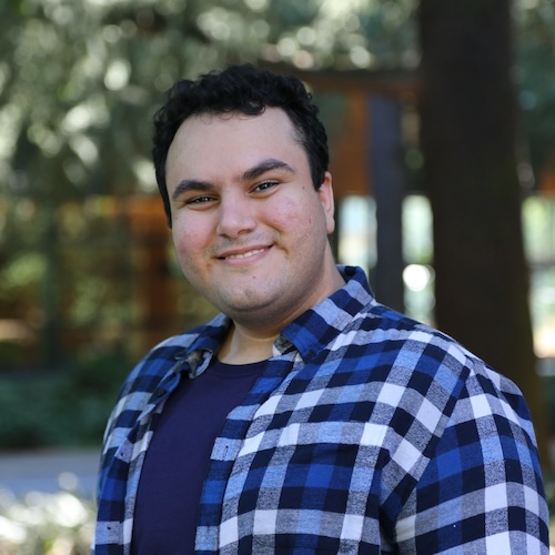 A profile picture of Ali Toghani, who is smiling in an outdoor setting. He is a white man with black hair wearing a blue and white plaid shirt over a dark t-shirt. The background features a natural environment, with trees.
