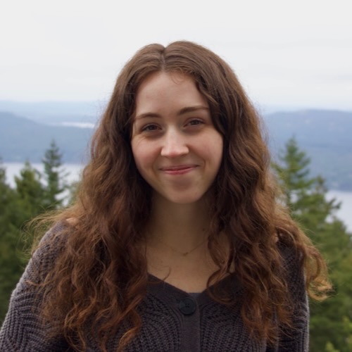 An image of Jaela, a white woman with brown curly hair wearing a grey sweater, smiling with evergreen trees, mountains, and ocean in the landscape behind her.
