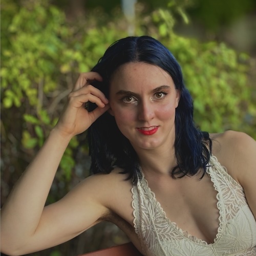 A profile picture of Katherine Leavitt smiling at the camera with some greenery in the background. She is a white woman with dark blue hair wearing a lacy white shirt. Her elbow is resting on a bench and she is touching her hair.
