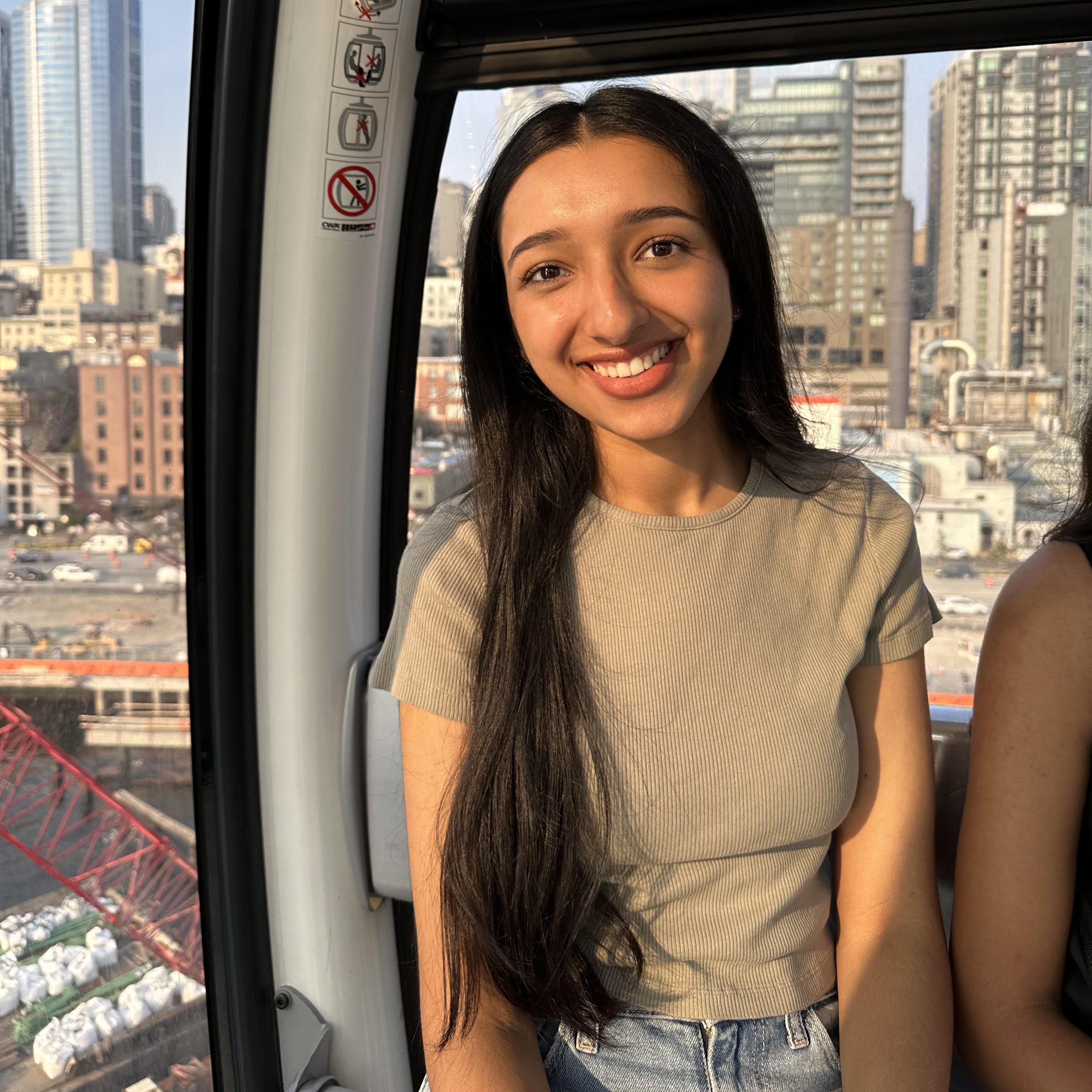 A profile picture of Anmol Bhuller, who is smiling at the camera with
     some Seattle buildings in the background. She has long black hair, brown skin, and is wearing
      a sage green T-shirt. 