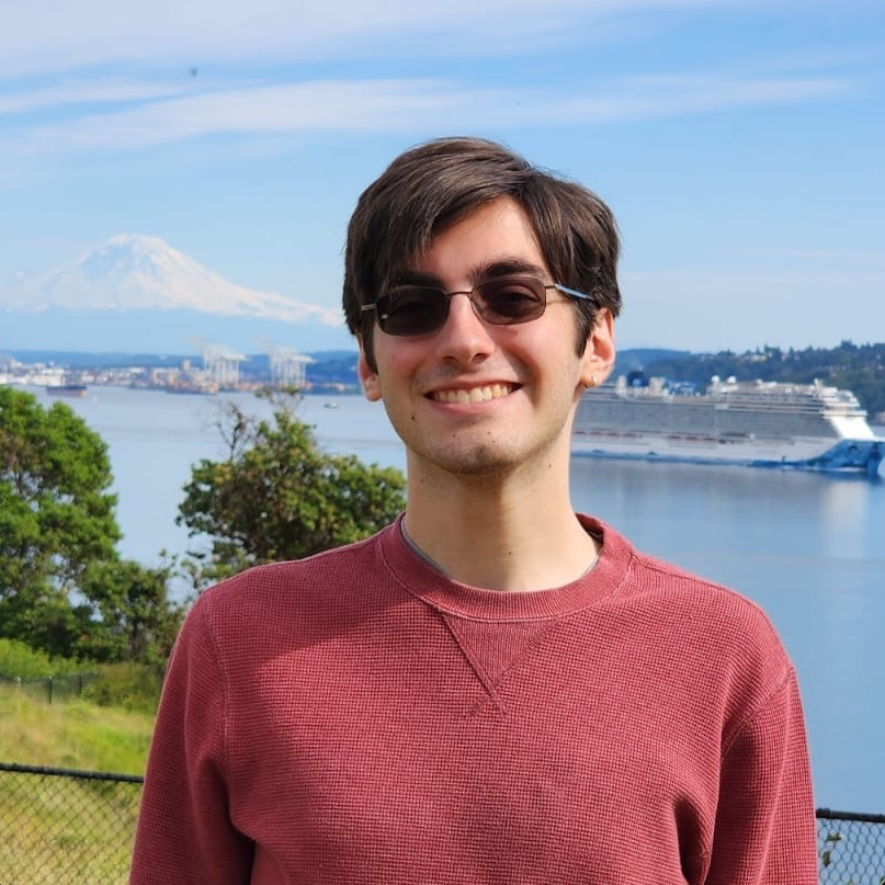 A profile photo of Antonio Ballesteros, who is smiling at the
    camera in front of the puget sounds with Mount Rainier in the background. He is a white
    man with brown hair, shaded glasses, wearing a red sweater.