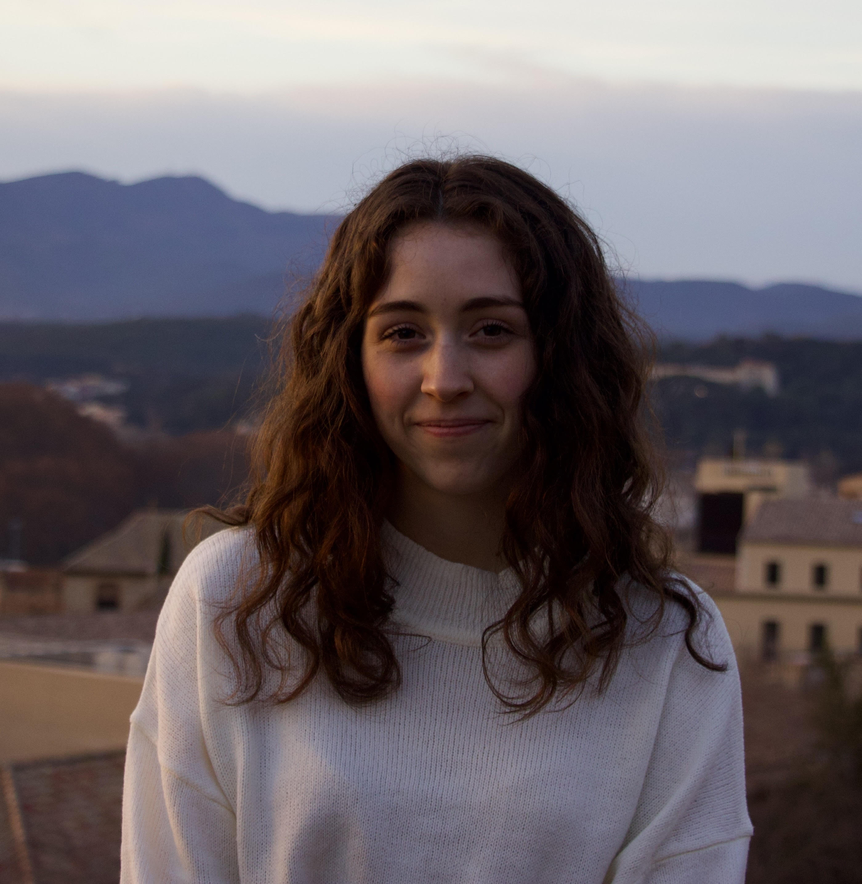 A profile picture of Jaela Field, who is smiling at the camera with
            tan buildings and blue-toned mountain in the background. She is a white woman with brown
            curly hair and a white sweater.