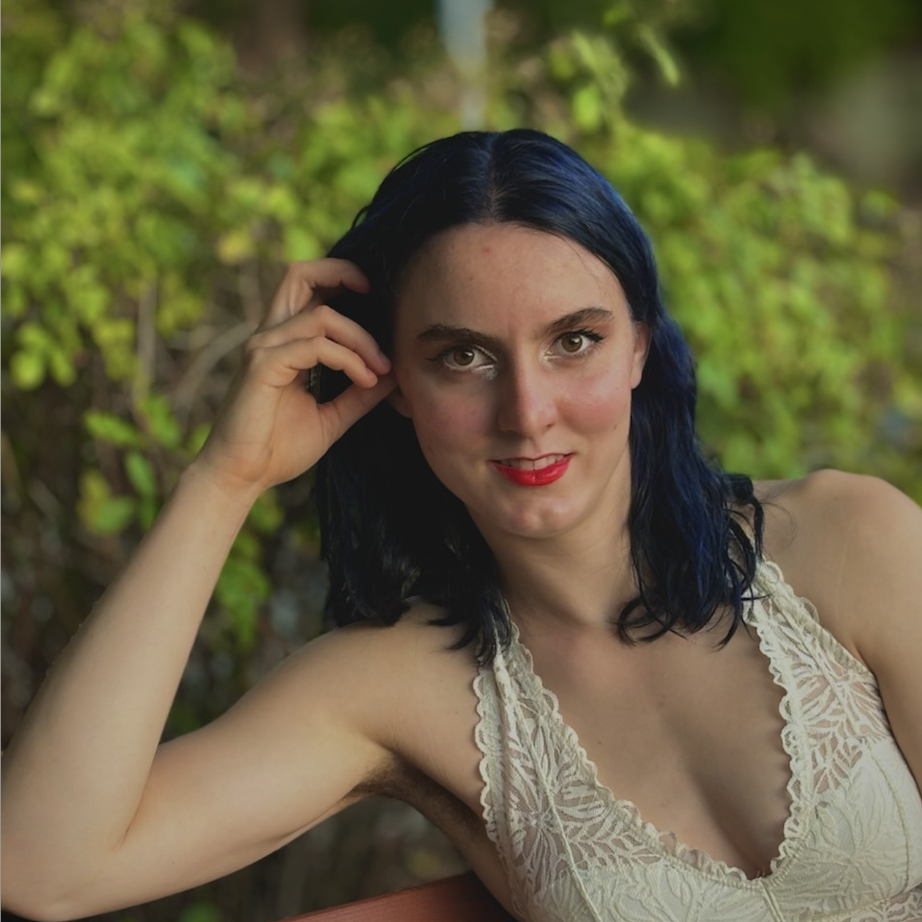 A profile picture of Katherine Leavitt smiling at the camera with
            some greenery in the background. She is a white woman with dark blue hair wearing a lacy
            white shirt. Her elbow is resting on a bench and she is touching her hair.