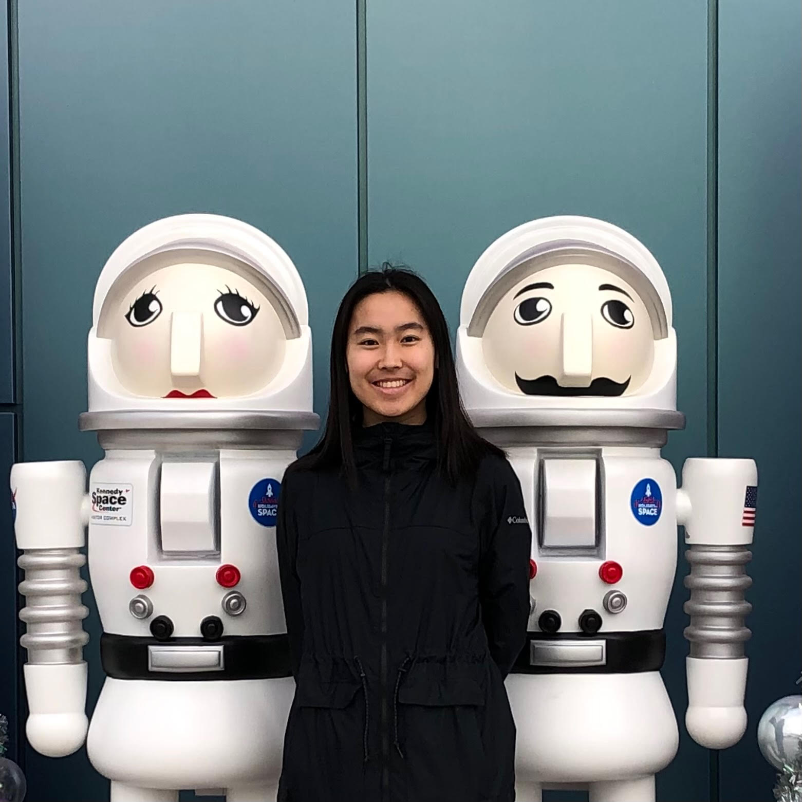 A picture of Mayee Sun between two nutcracker statues 
    dressed in astronaut suits. She is an Asian woman wearing a black rain jacket and 
    standing with her arms behind her back.