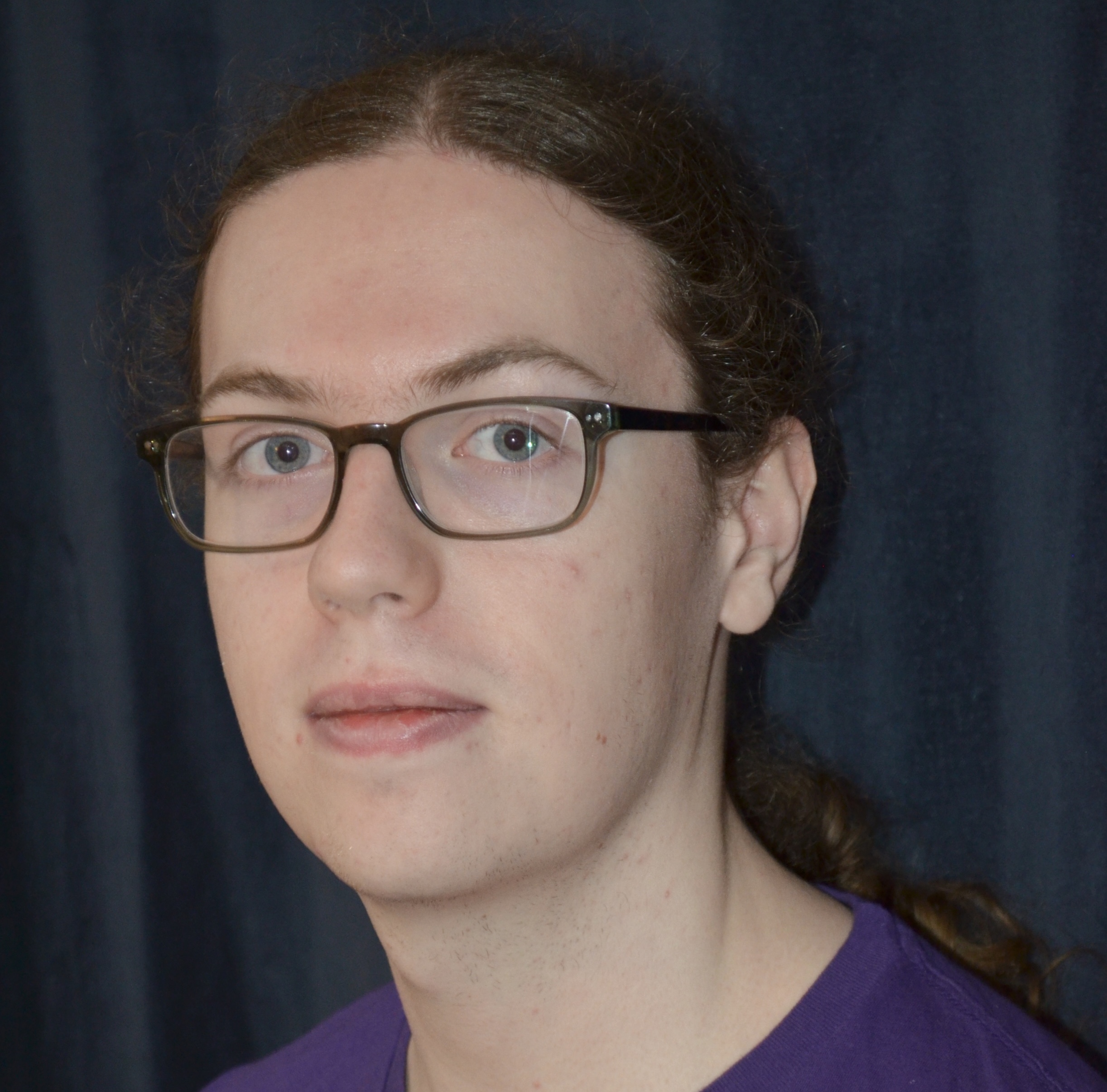 A profile picture of Noah Huck, who is standing against a dark 
    grey background and smiling at the camera. He is a white man with long brown hair, wearing 
    glasses and a purple T-shirt.