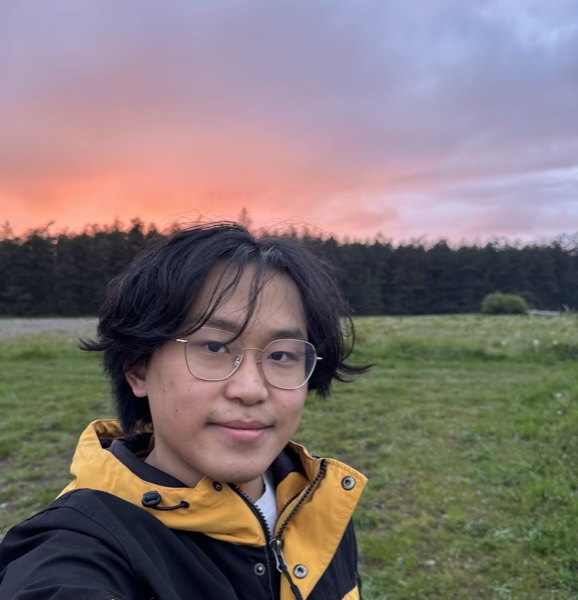 Owen in glasses and a yellow and black jacket, in a grassy field under a pink sunset sky.