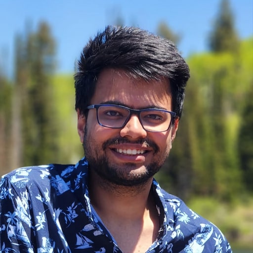 Reshabh with glasses in a patterned blue shirt smiles in an outdoor setting with trees in the background.