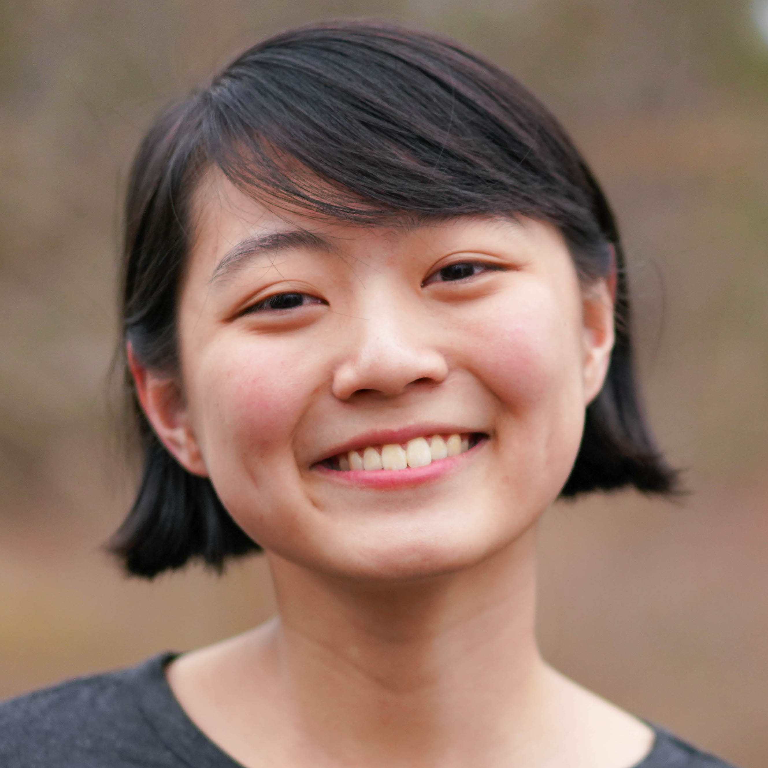 A headshot photo of Audrey. They are wearing a gray shirt and are smiling. The background is brown and blurry.