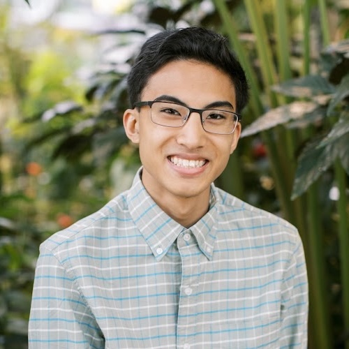 A profile picture of Raio, sitting outdoors, wearing a plaid shirt, and smiling.