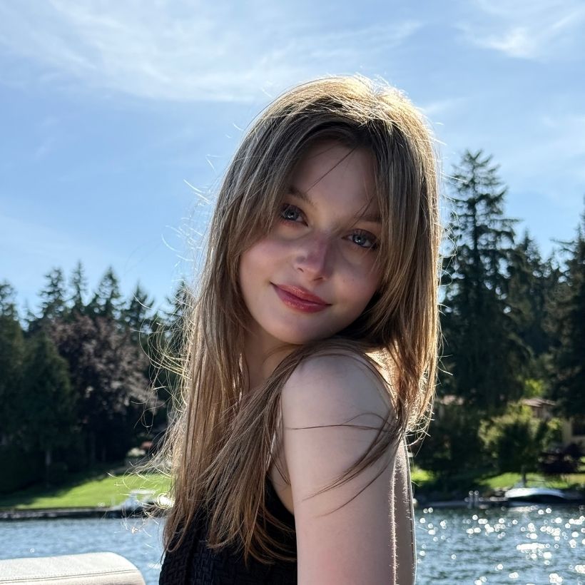A profile picture of Saan sitting outdoors with water and trees in the background. She is wearing a black shirt and smiling.
