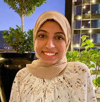 Headshot of Fatimah, who is standing in front of a tall skyscraper and a garden with trees.