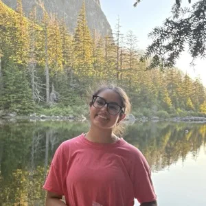 Picture of Aashi Juneja (she/her). Her photo shows her smiling outside with trees in the background.