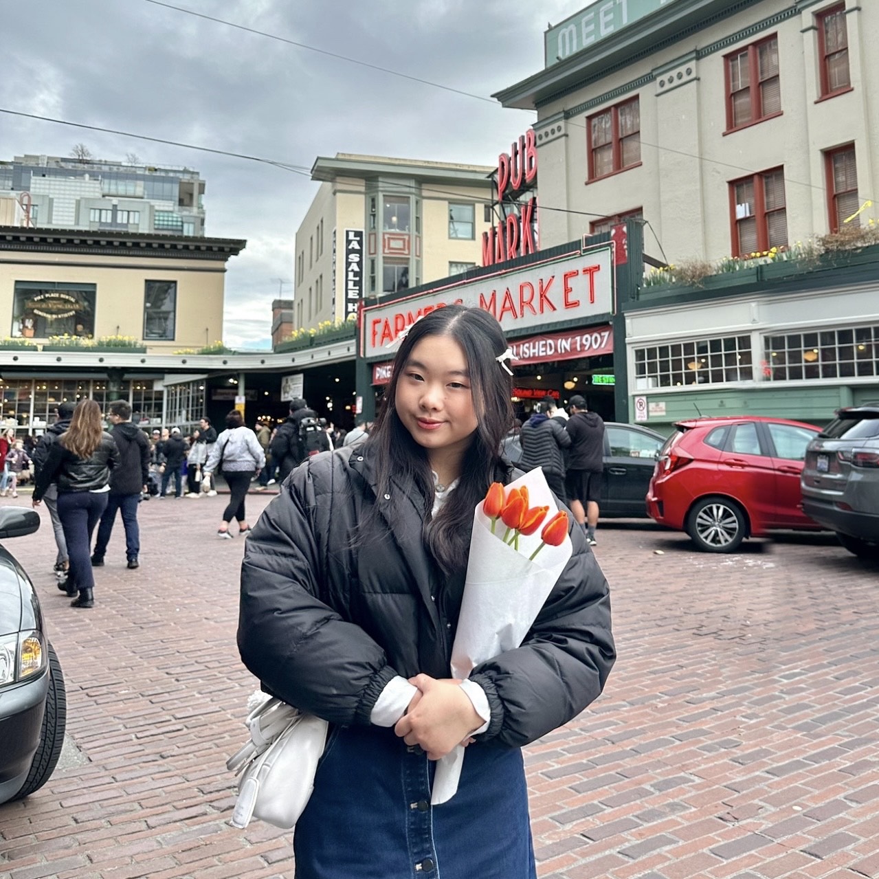 Megan Wangsawijaya smiling and holding a bouquet of five orange tulips at Pike Place Market. She is wearing a black puffer jacket, denim skirt, and a white crossbody bag.