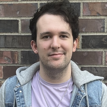 A light-skinned, masc-presenting person with short, wavy dark hair, wearing a pink t-shirt under a denim hoodie, with a steel necklace, and eyebrow and ear piercings.