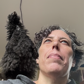 a headshot of a white person with short curly grey-streaked her. A chicken sits on their shoulder.