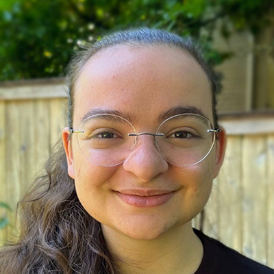 A headshot of Naama smiling at the camera. She is a white
    woman with long brown hair and glasses. She is wearing a black T-shirt.