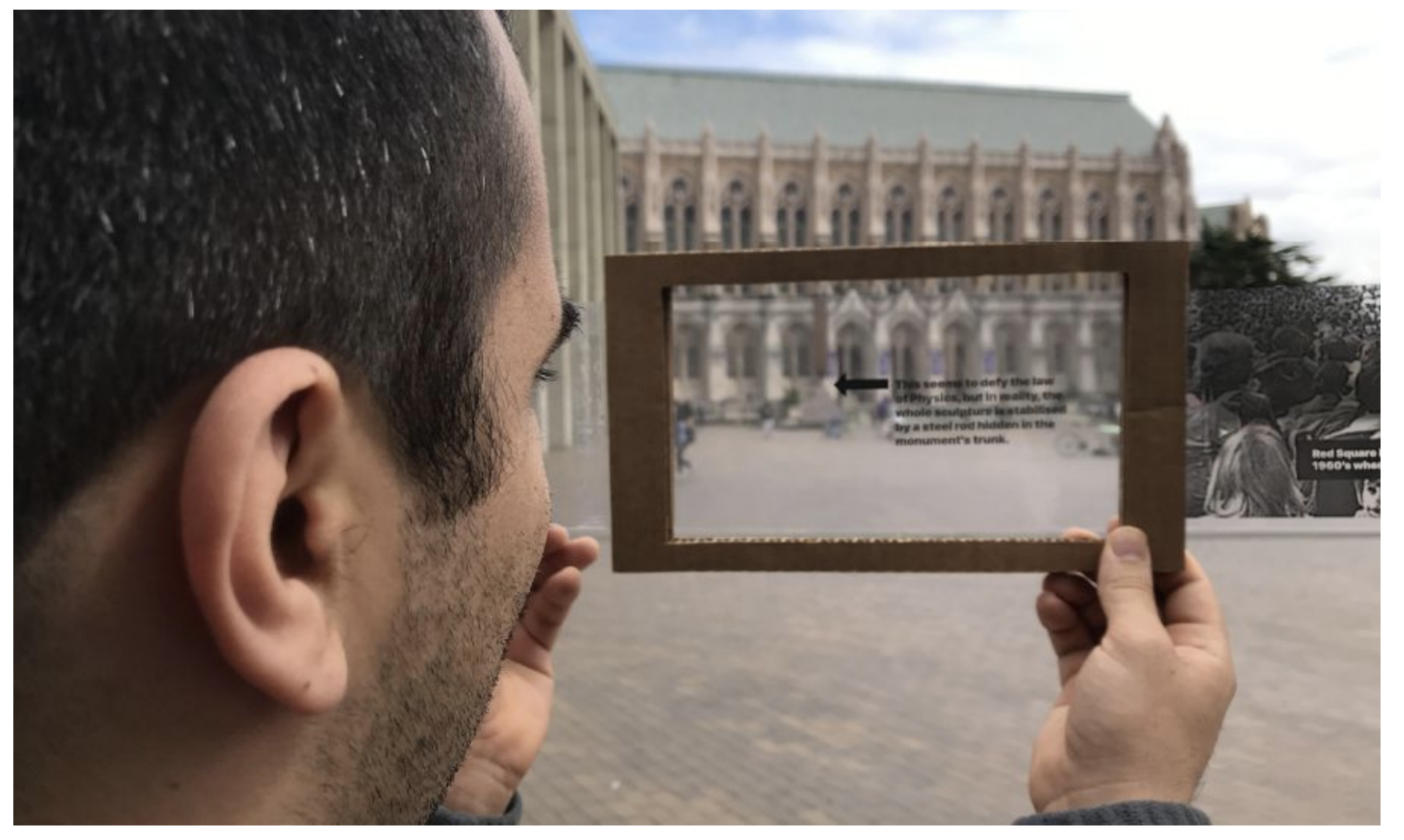 A closeup of a person looking through a see-through screen made of cardboard. An arrow and text on the screen are pointing at a building in the background