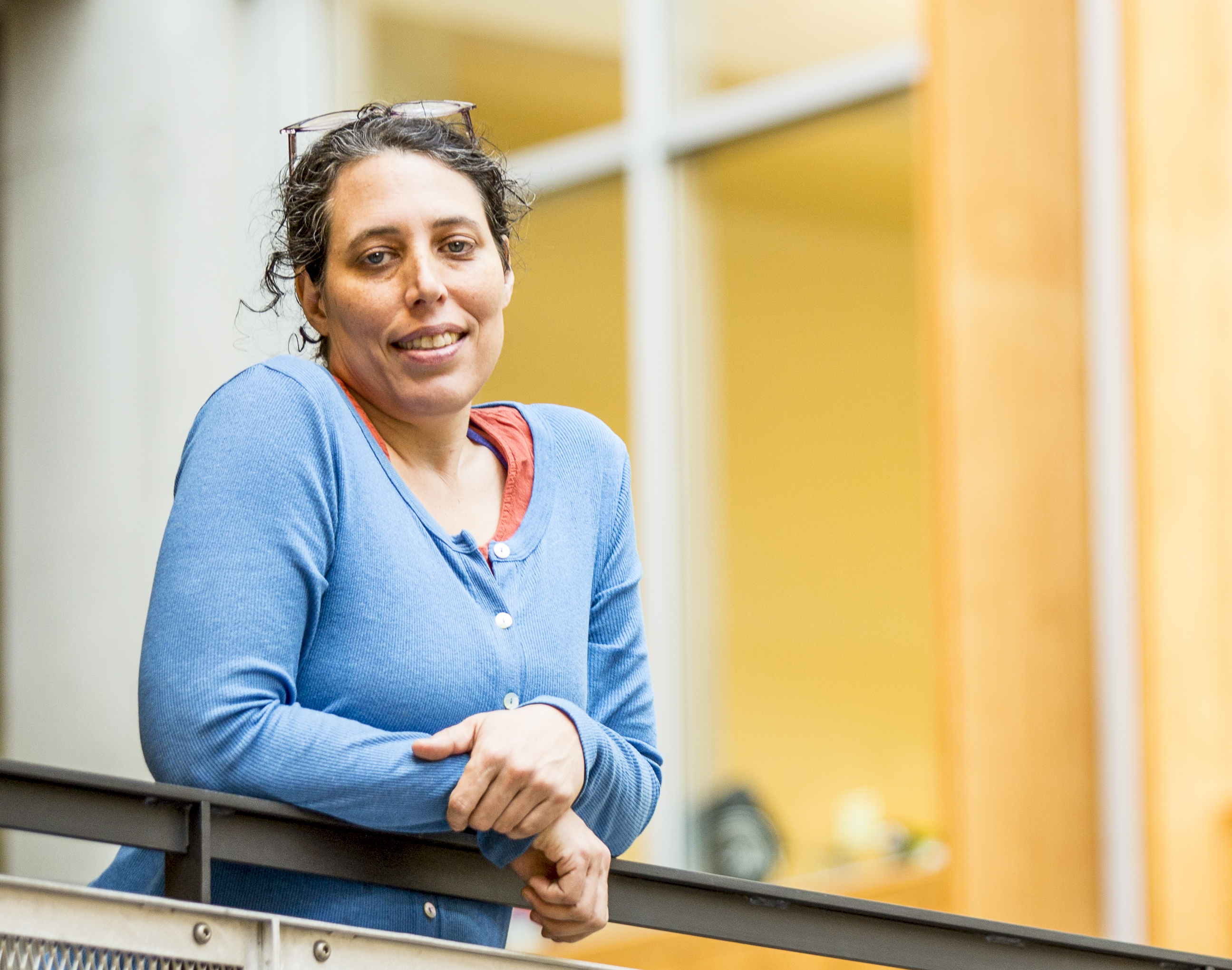 Photograph of a white woman with curly brown hair and glasses on her head in a blue shirt learning on a railing
