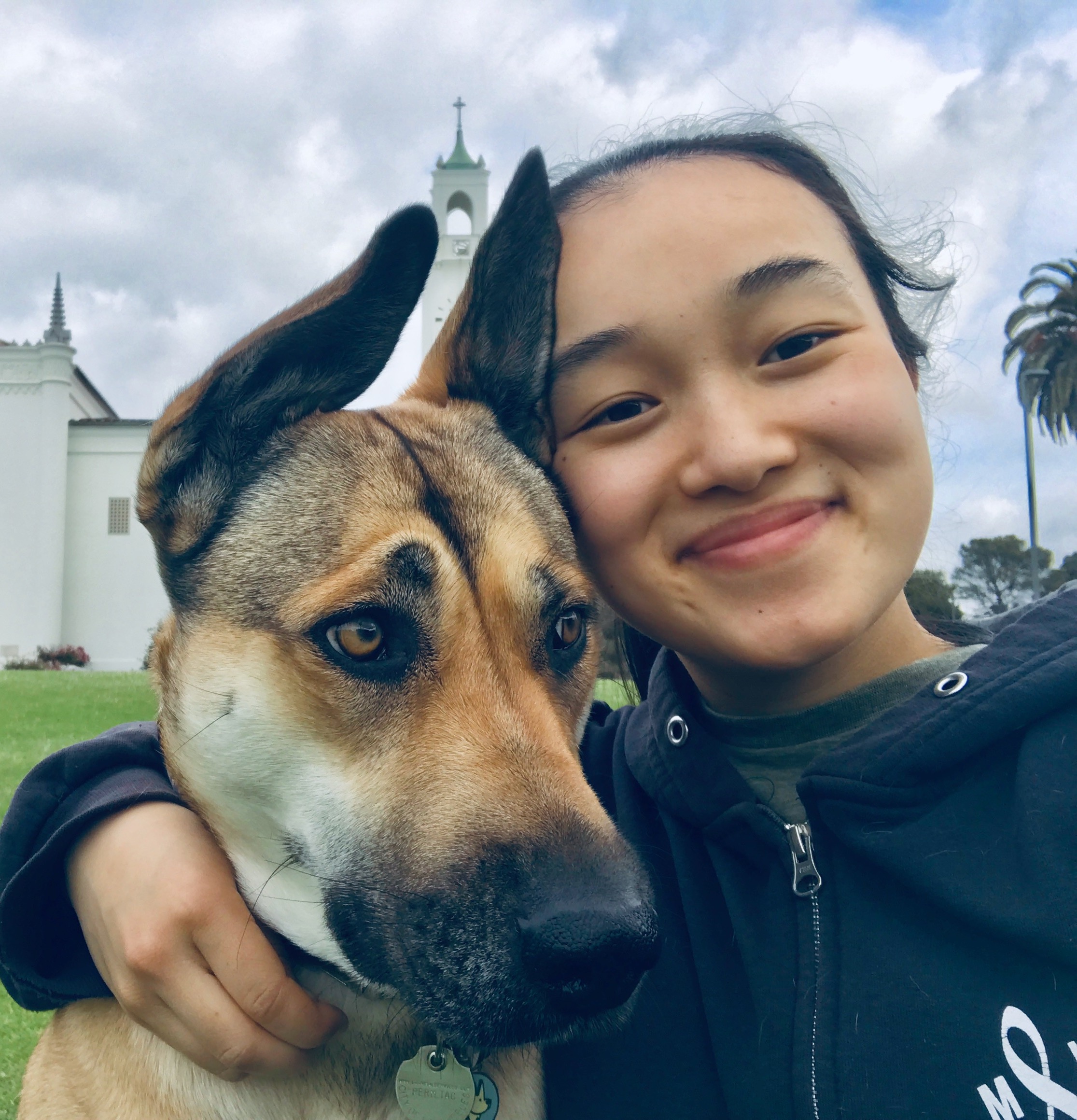 A selfie of an Asian woman with black hair tied back in a ponytail, hugging a dog with tan and black fur and crooked ears, on an open field.