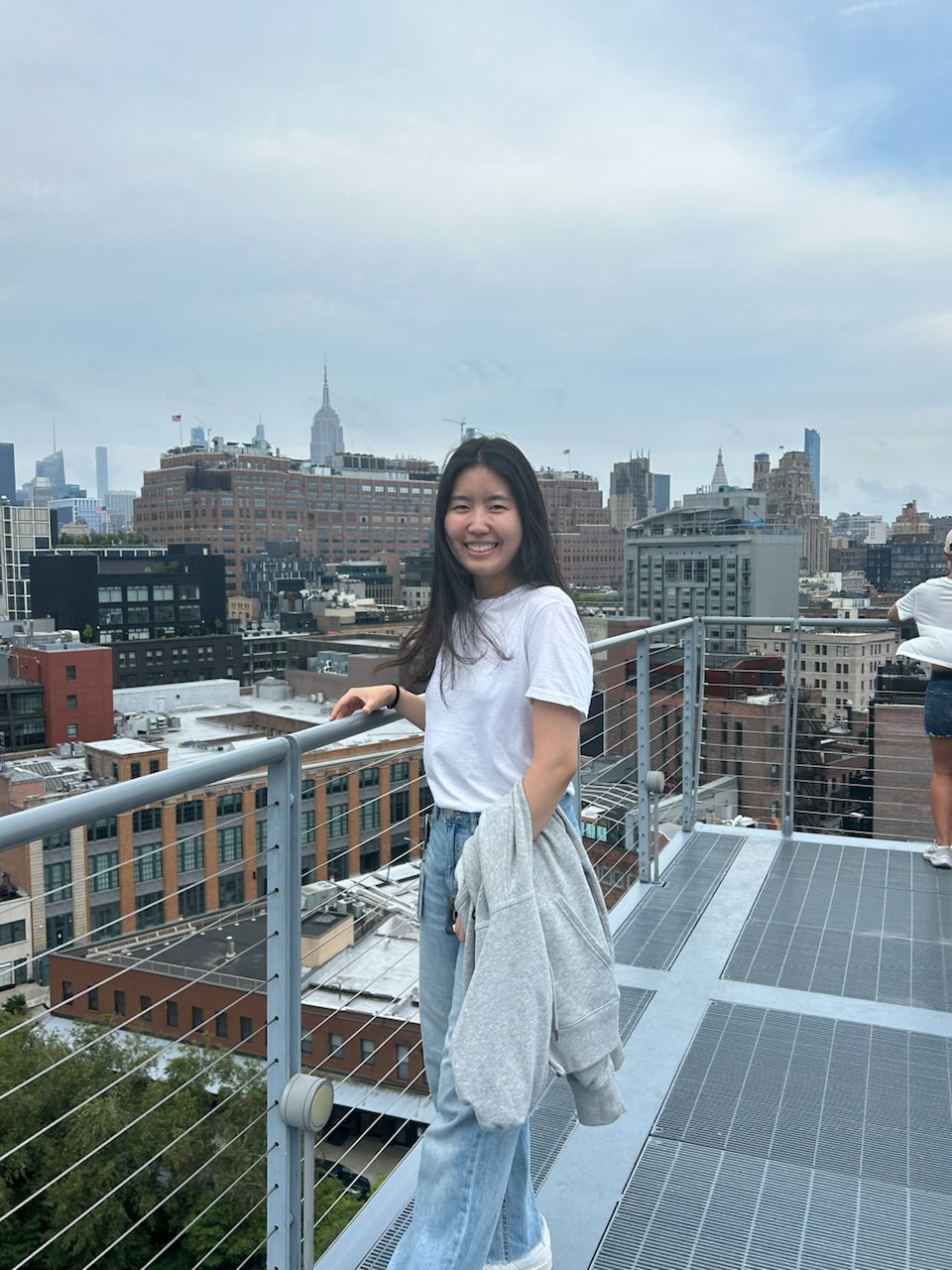 A picture of a young Asian adult female with long dark hair who is standing in front of a fence of a rooftop, wearing a white t-shirt and light-blue jean