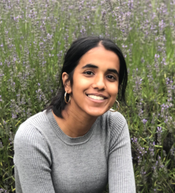 Photograph of a Indian woman with straight black hair in a ponytail and in a gray sweater surrounded by a field of lavenders