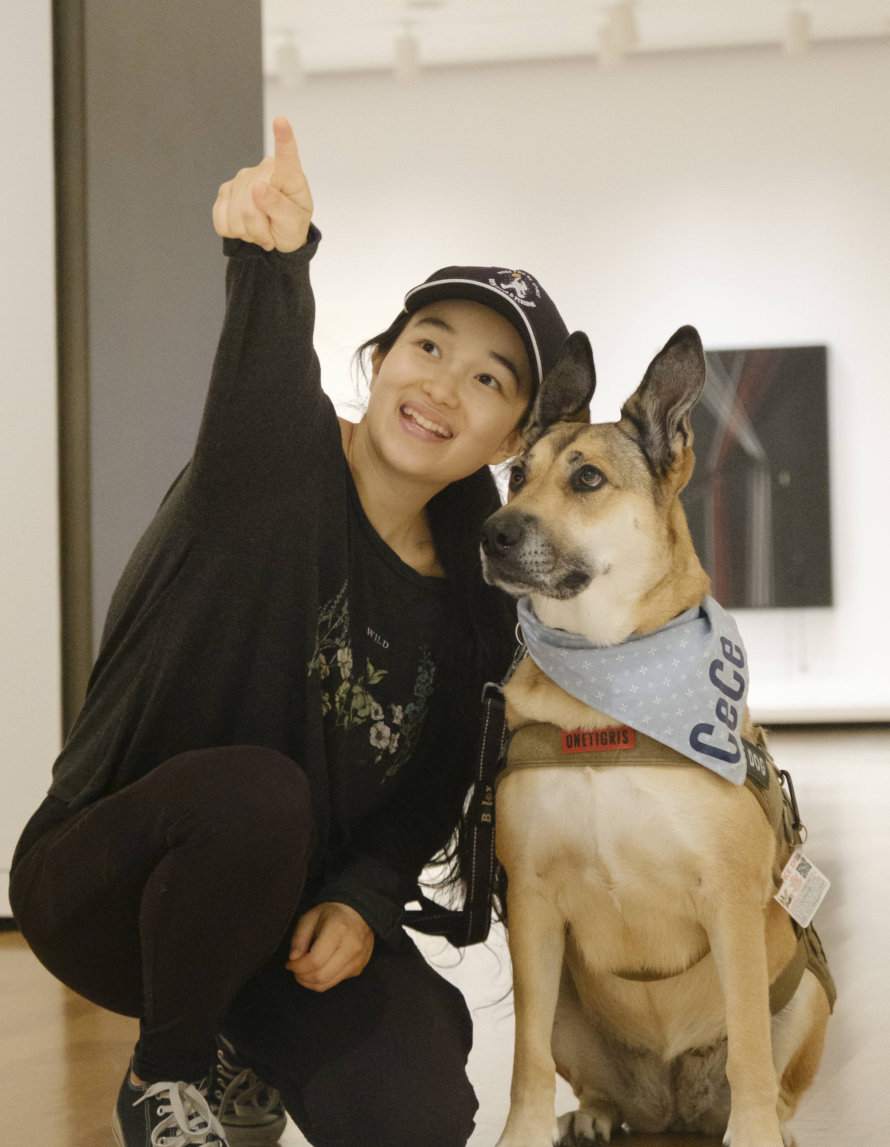 A picture of an Asian woman with a black ponytail pointing to something off screen and kneeling on the ground next to a tan-and-black service dog with crooked ears